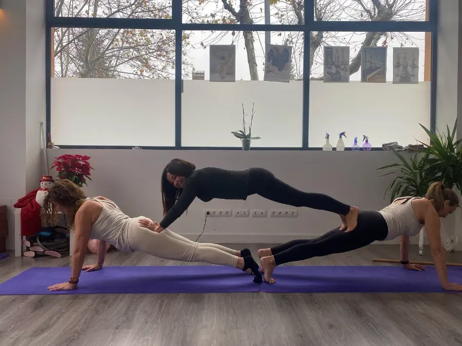Tres personas practicando la postura de la plancha en un estudio de yoga. Una persona está apilada sobre otras dos.