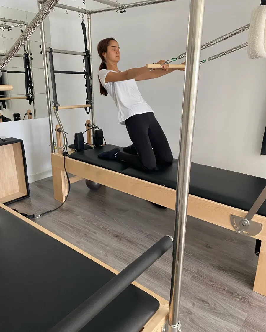 Mujer practicando Pilates en un reformer, tirando de las correas de rodillas. Habitación de color claro.