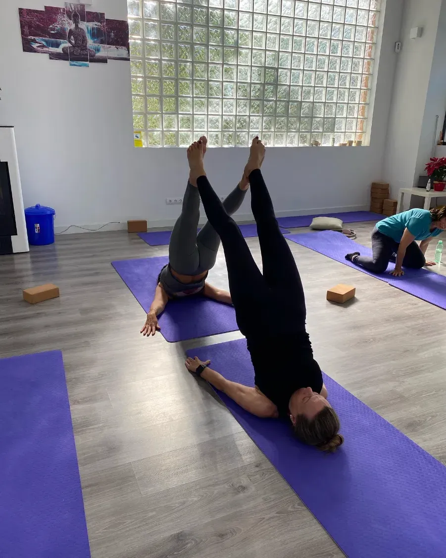 Mujer en un reformador de pilates, con los brazos por encima de la cabeza, haciendo split. Ropa deportiva negra, estudio interior.
