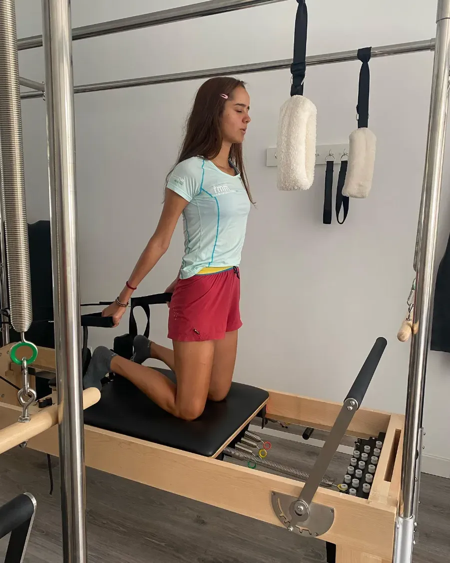 Mujer en un reformador de Pilates, de rodillas, tirando de las asas. Vestida con ropa deportiva en un estudio.