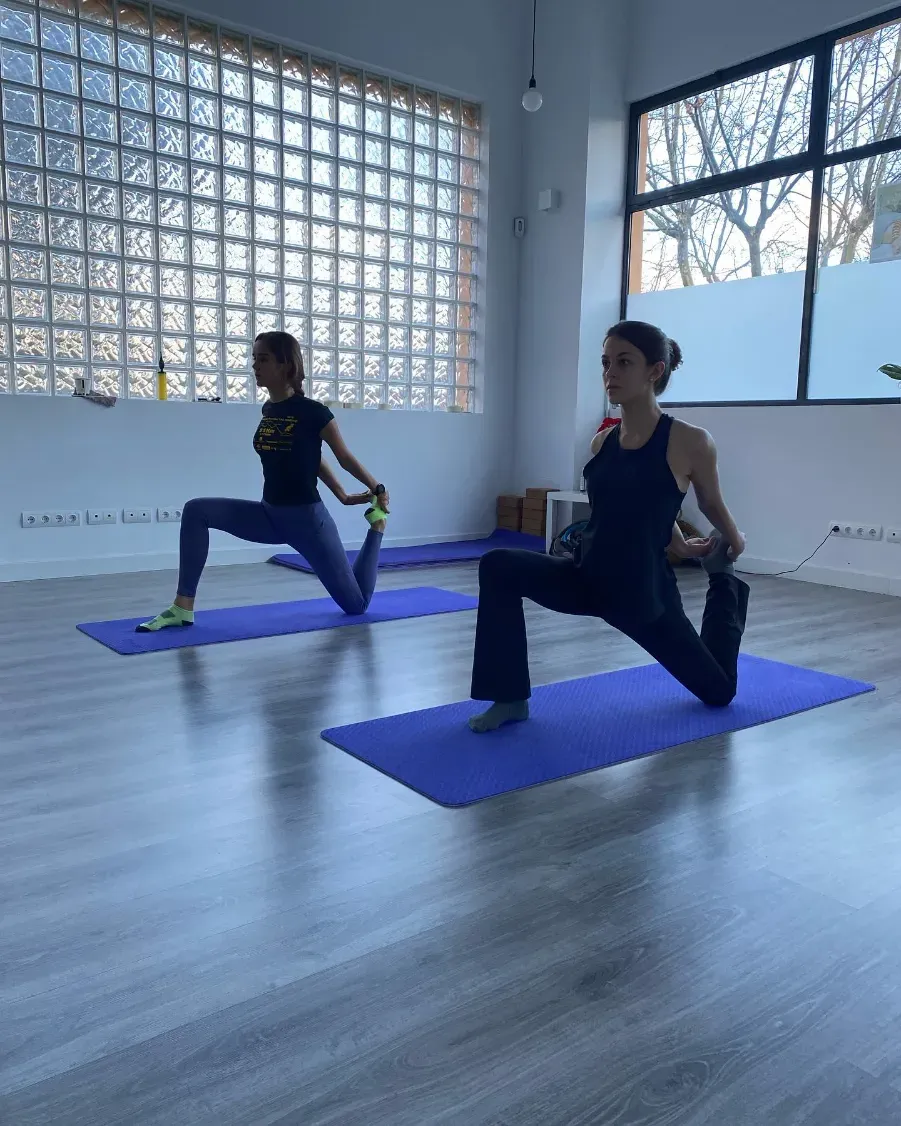Dos personas haciendo estiramientos de yoga sobre colchonetas azules en un estudio con ventanas.