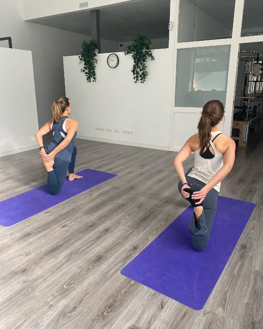 Dos mujeres en un estudio de yoga, con el pie apoyado sobre esterillas moradas. Paredes blancas, suelo gris.