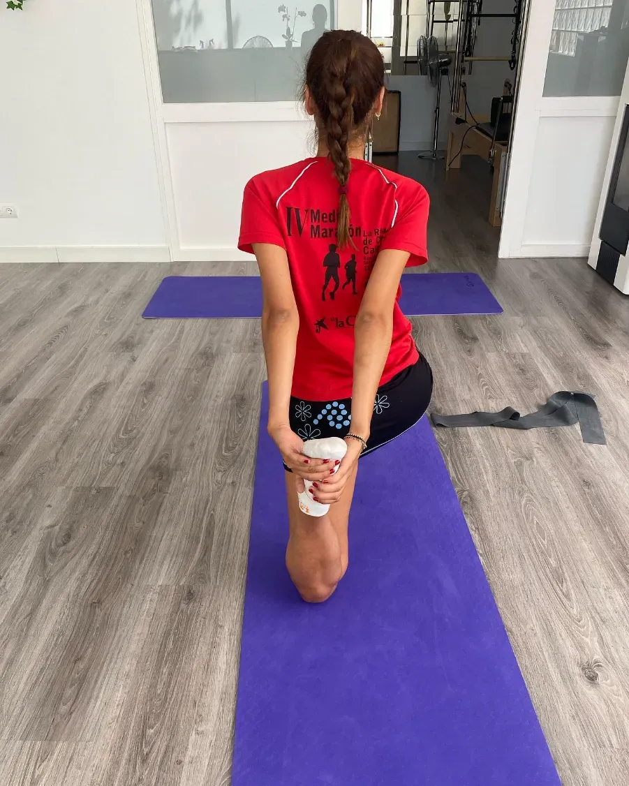 Mujer con camisa roja haciendo una pose de yoga sobre una colchoneta morada, sosteniendo un pie detrás de ella.