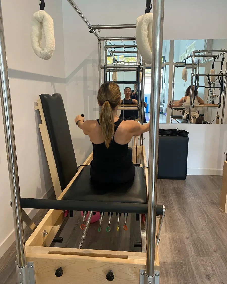 Mujer vestida con ropa deportiva negra en una máquina de pilates, con los brazos extendidos, en un estudio con espejos.