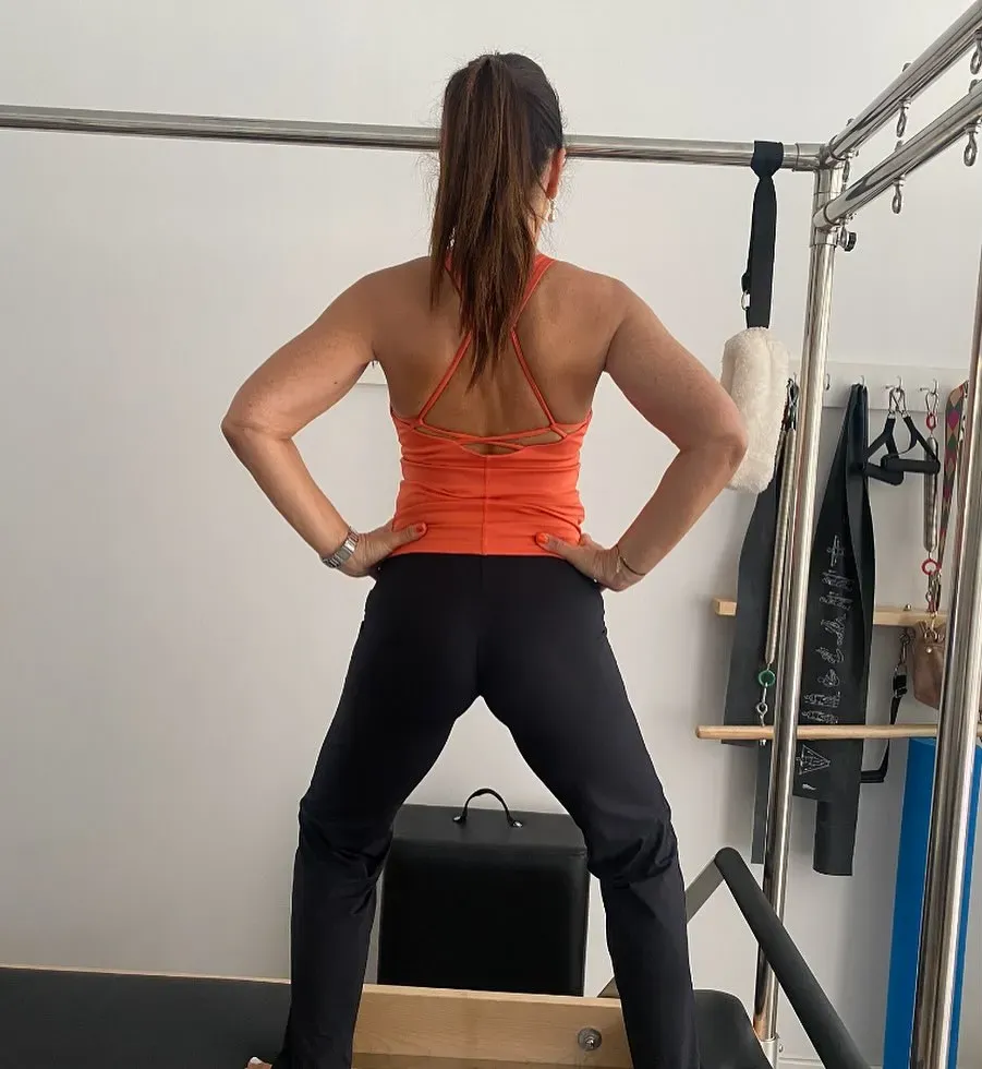 Mujer con camiseta naranja y pantalón negro, con las manos en las caderas, de pie sobre un reformador de Pilates.
