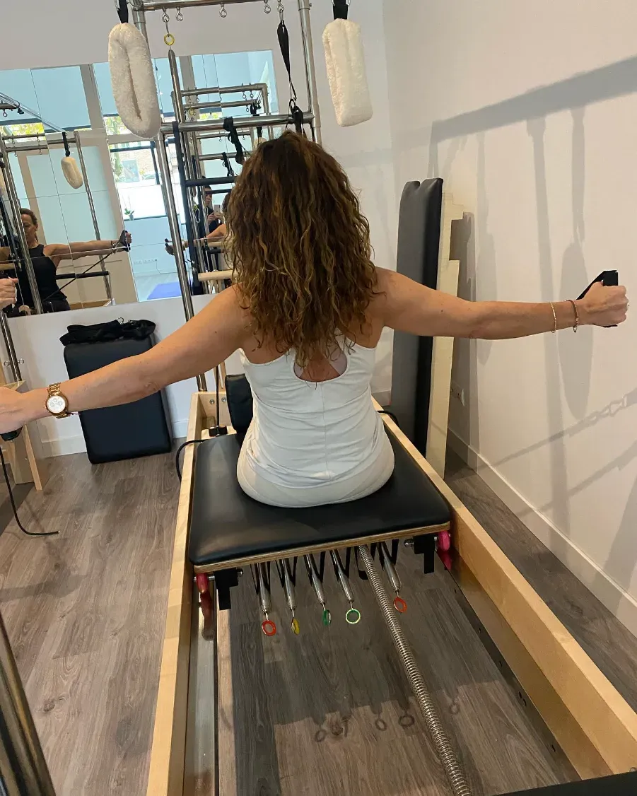 Mujer sentada en un reformer de Pilates, con los brazos extendidos a los lados, sujetando las asas. Interior.