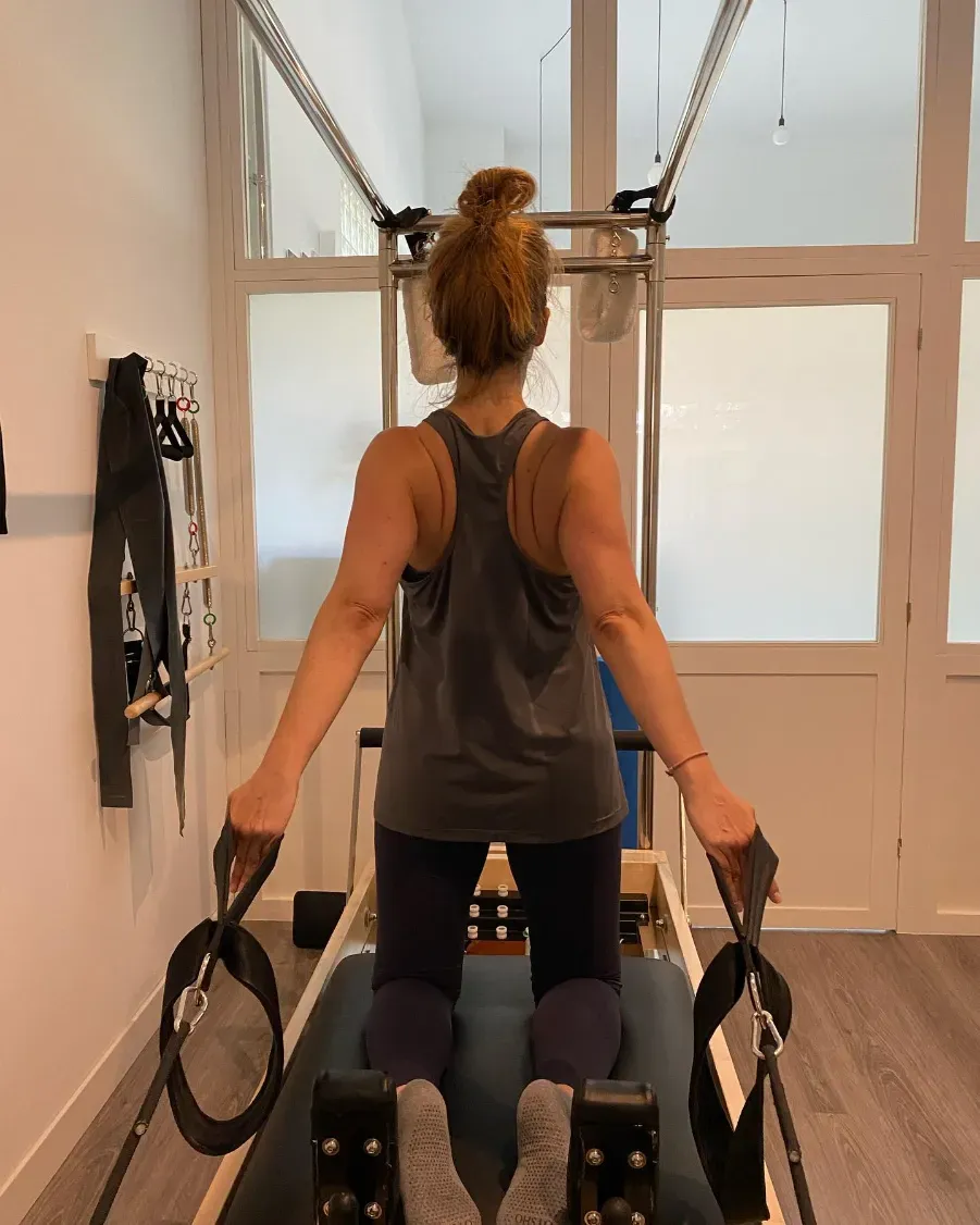 Persona arrodillada sobre un reformador de Pilates, sujetando correas, haciendo ejercicio en un estudio.