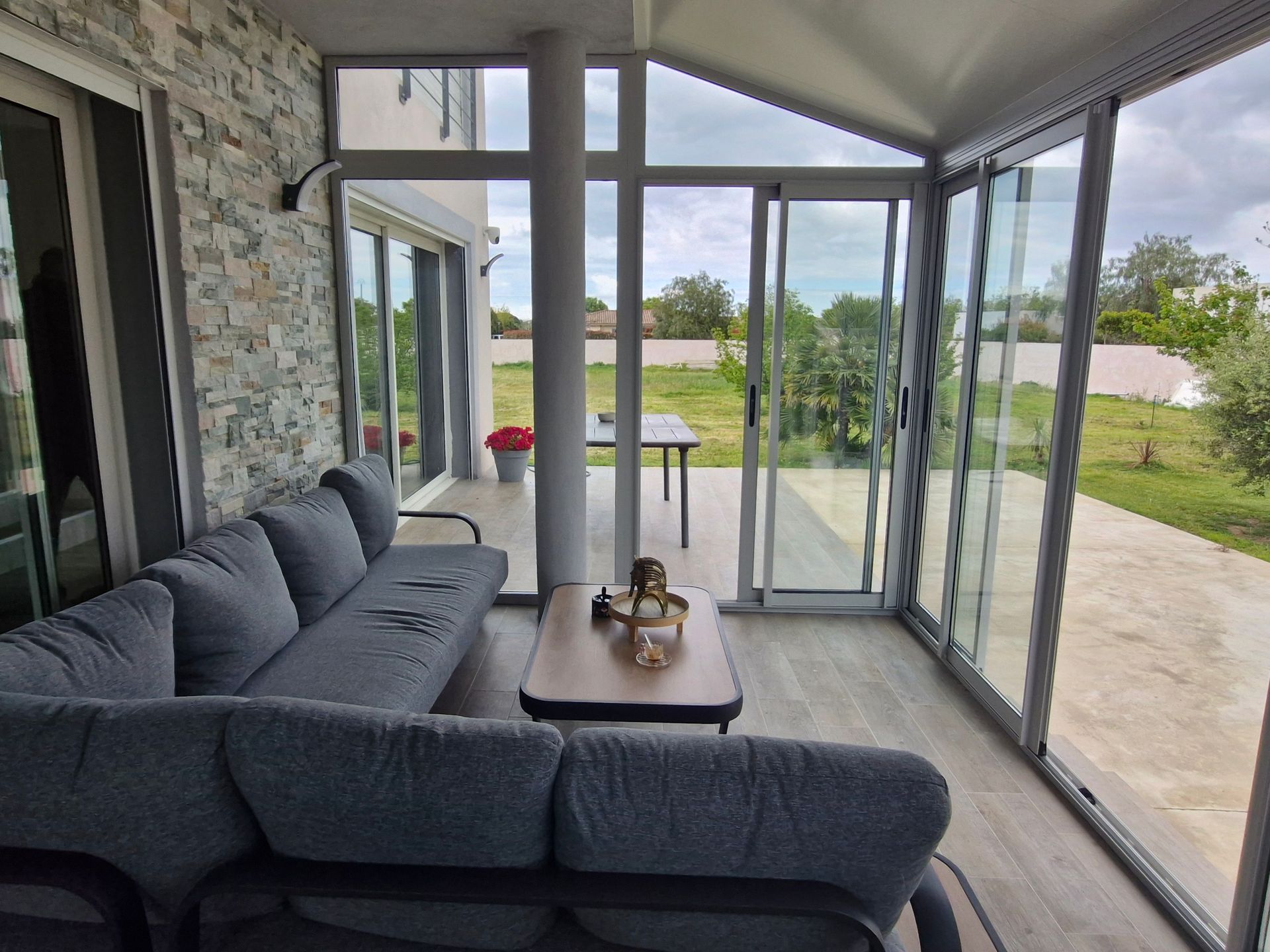 Un coin salon extérieur confortable avec un canapé d'angle gris, des parois vitrées et une vue sur un jardin
