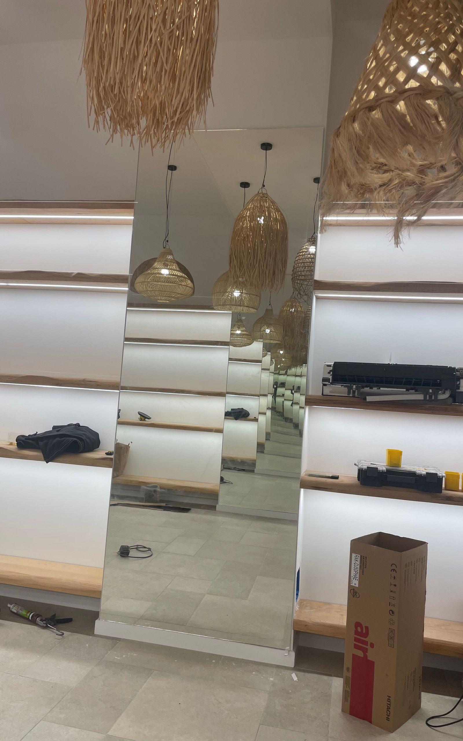 Un agencement de magasin moderne avec des étagères en bois clair, des luminaires suspendus texturés et un grand miroir central sur pied.