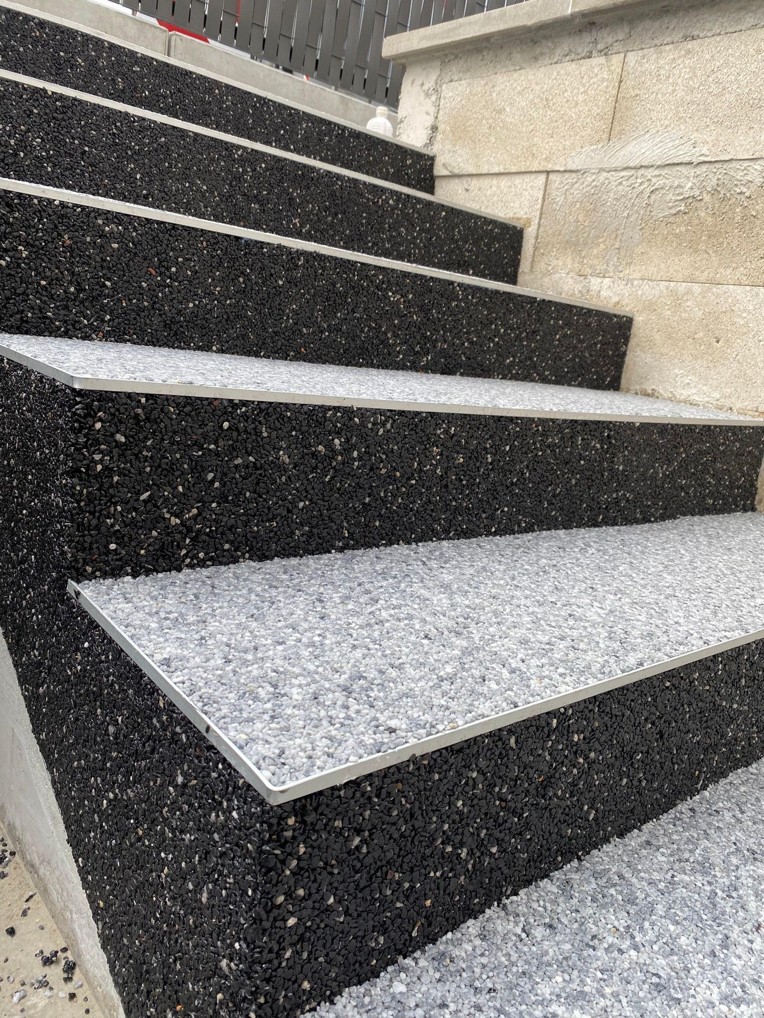 Escalier extérieur avec finition mouchetée noire et grise, bordure en métal et mur en pierre.