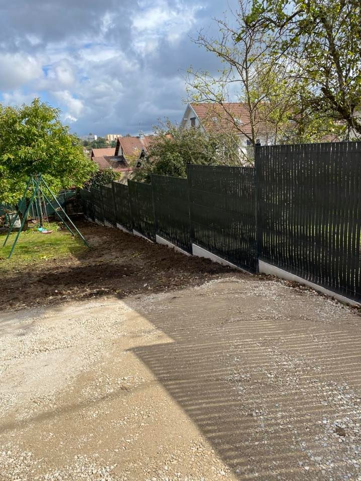 Clôture sombre à côté d'une allée de gravier, menant à une cour verte et des arbres sous un ciel nuageux.