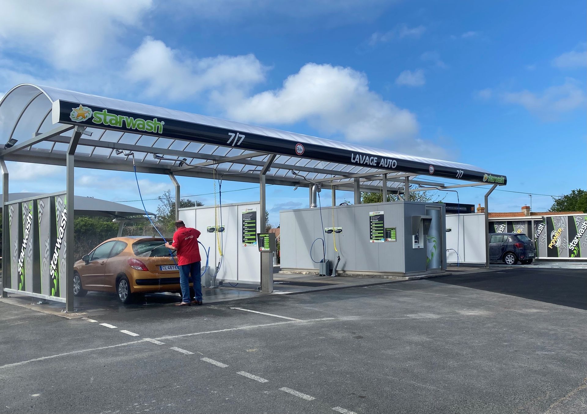 Station de lavage auto avec une voiture orange, une personne la lavant. Extérieur sous un ciel bleu.