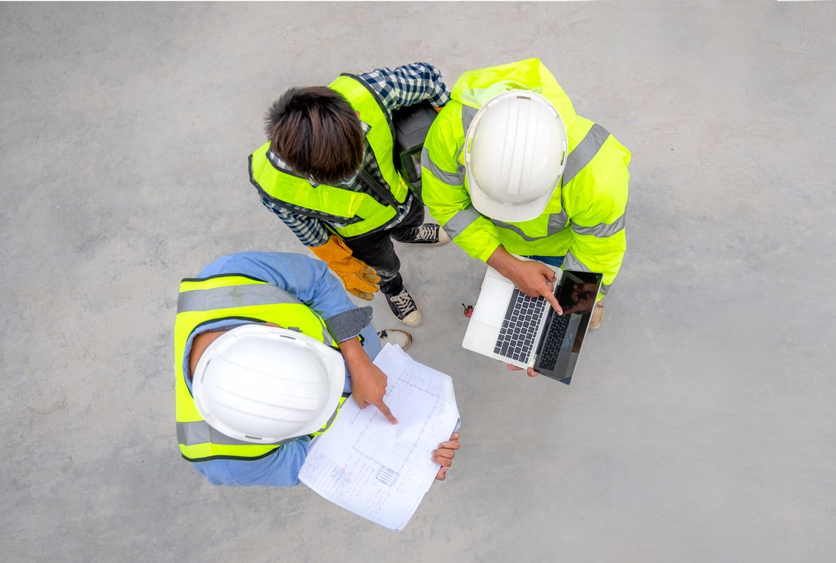 Trois ouvriers du bâtiment, portant des gilets de sécurité et des casques de chantier, examinent des plans et un ordinateur portable.