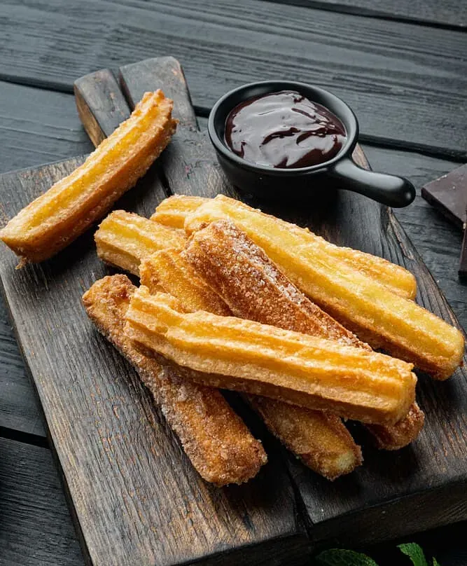 Una tabla de cortar de madera cubierta con churros y un tazón de salsa de chocolate.