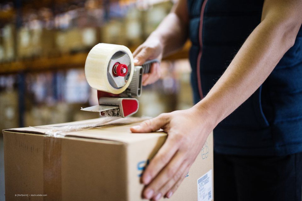 GUNTHER BLUM GmbH – eine Fachkraft in Warnweste und mit Handschuhen trägt einen Karton zu einem Kartonstapel in einem Lager