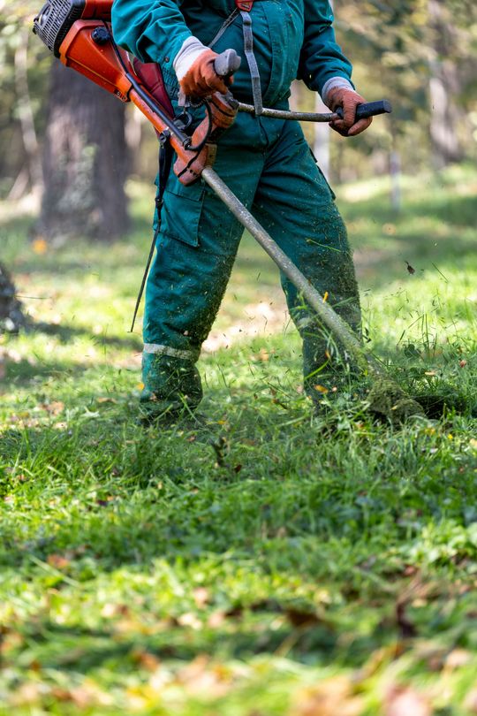 Une personne en salopette verte utilise une débroussailleuse à gazon pour couper de l'herbe à l'extérieur.