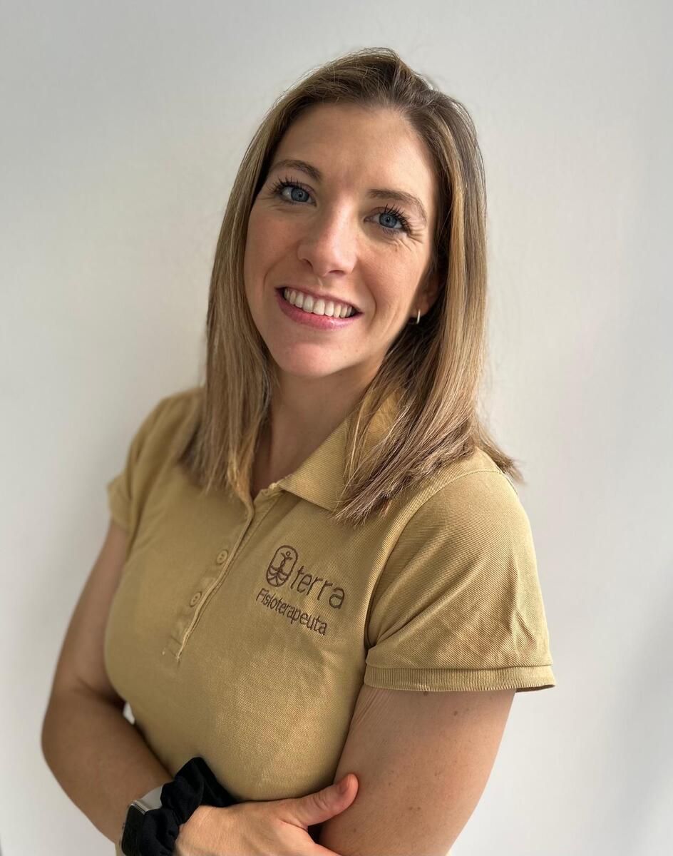 Mujer de cabello rubio sonríe, vistiendo un polo color canela con logo; con los brazos cruzados.