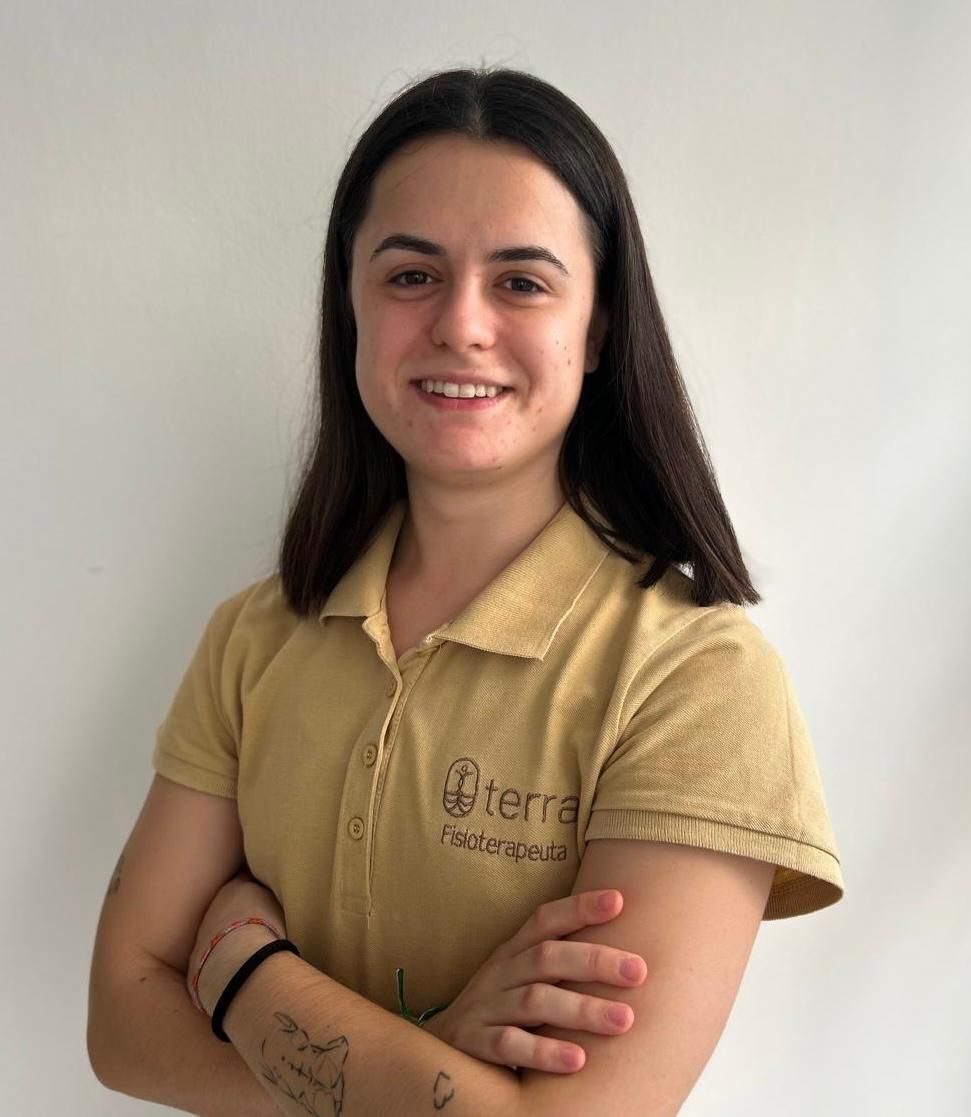 Una mujer de cabello oscuro y un polo color canela sonríe, con los brazos cruzados, frente a una pared blanca.