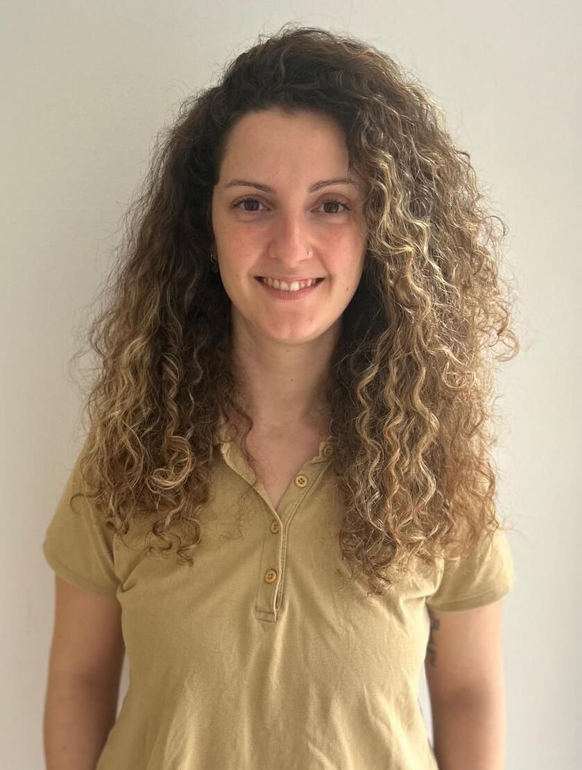 Una mujer de cabello castaño y rizado sonríe, vistiendo una camisa color canela, contra una pared blanca.