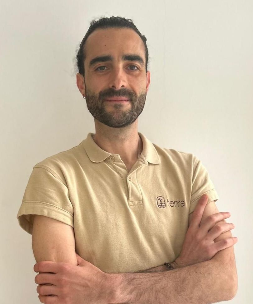 Hombre de cabello oscuro con un polo color canela, con los brazos cruzados, mirando a la cámara. La camiseta tiene un logo.