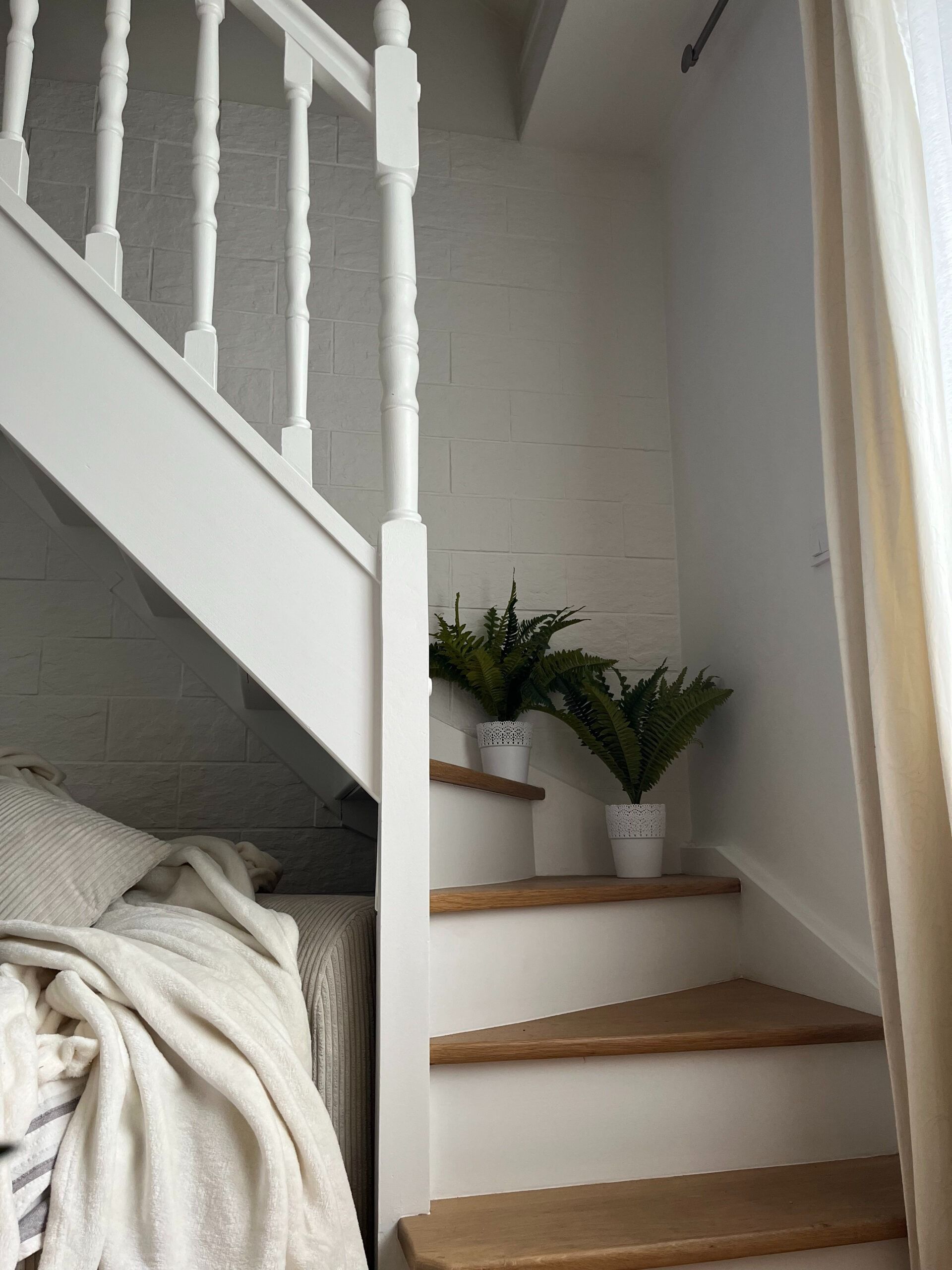 Escalier blanc aux marches en bois, deux fougères en pot sur le palier, mur de briques blanches.