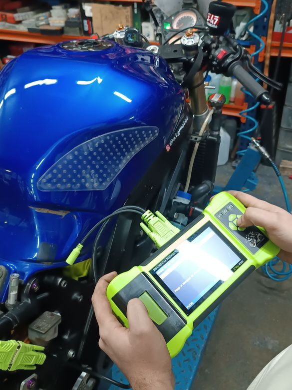Manos sosteniendo un dispositivo de diagnóstico amarillo frente a una motocicleta azul en un taller.