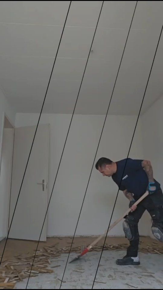 Une personne en tenue de travail utilise un grattoir à sol pour enlever des résidus du sol d'une pièce.