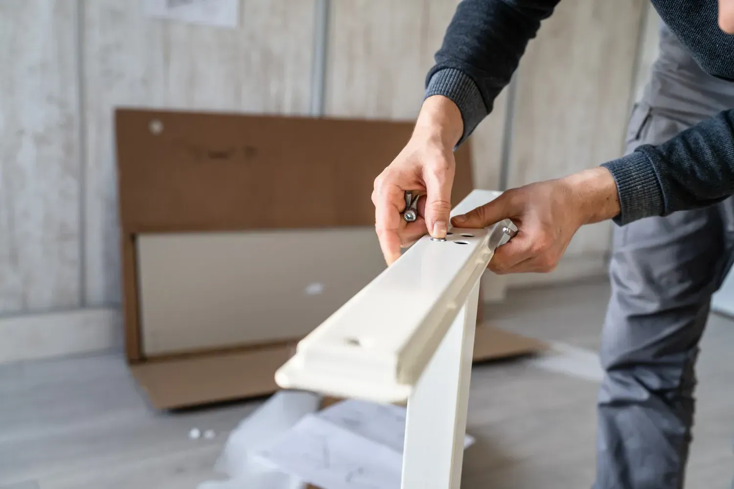 Persona ensamblando muebles blancos, sosteniendo piezas y una herramienta, caja de cartón en el fondo.
