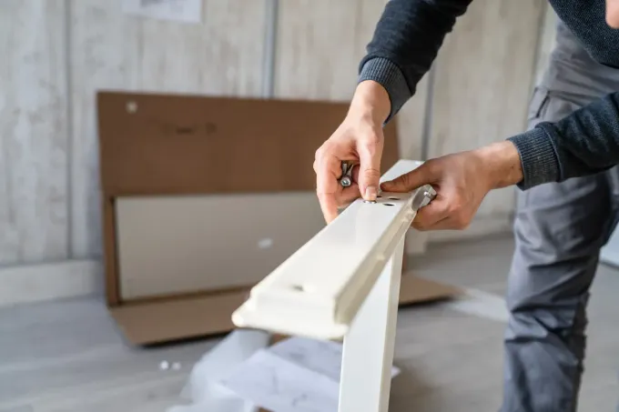 Persona ensamblando muebles blancos, sosteniendo piezas y una herramienta, caja de cartón en el fondo.