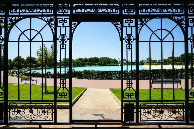 Una puerta de hierro negro adornada enmarca una vista de un parque con un estanque turquesa y un cielo azul.
