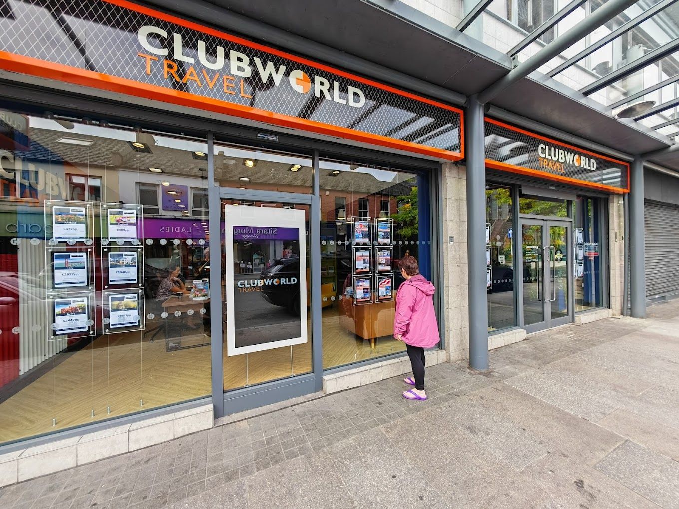 A woman in a pink jacket is standing in front of a clubworld travel store.