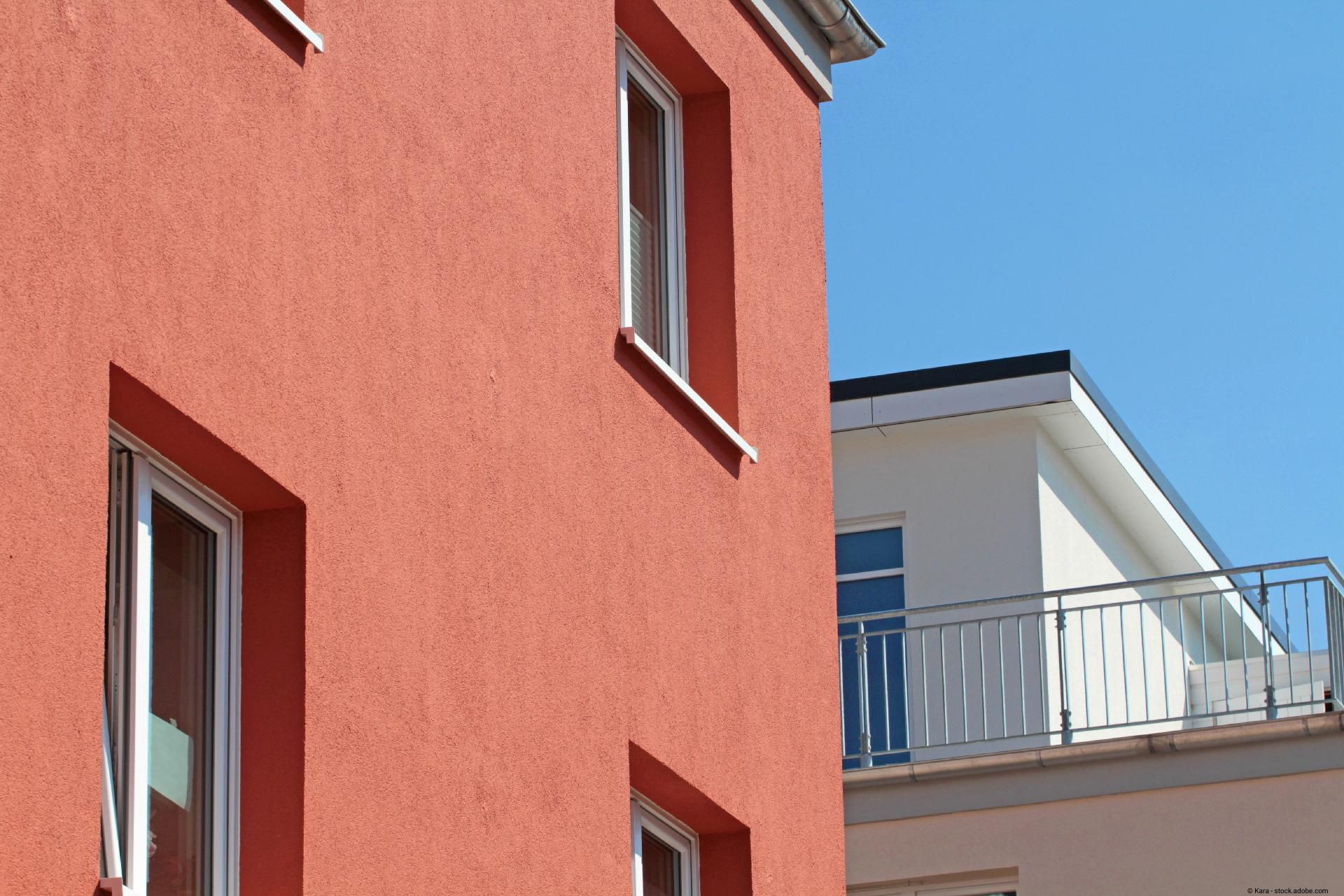 Rotbraun verputzte Hausfassade mit Fenstern im Vordergrund und ein weißes Gebäude.