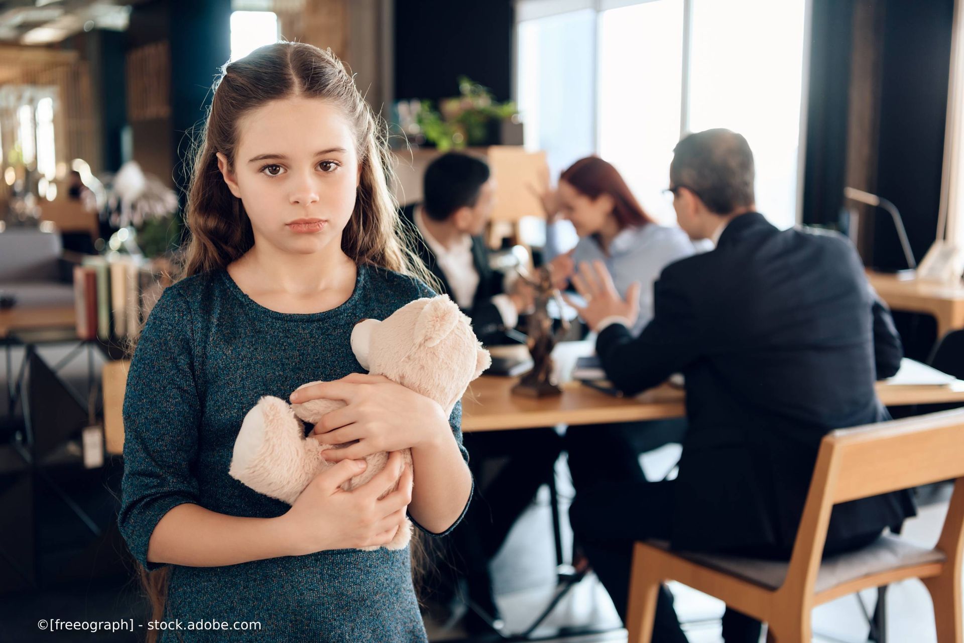 Ein kleines Mädchen hält vor einer Gruppe von Menschen einen Teddybären. Rechtsanwaltskanzlei UDO SAUL