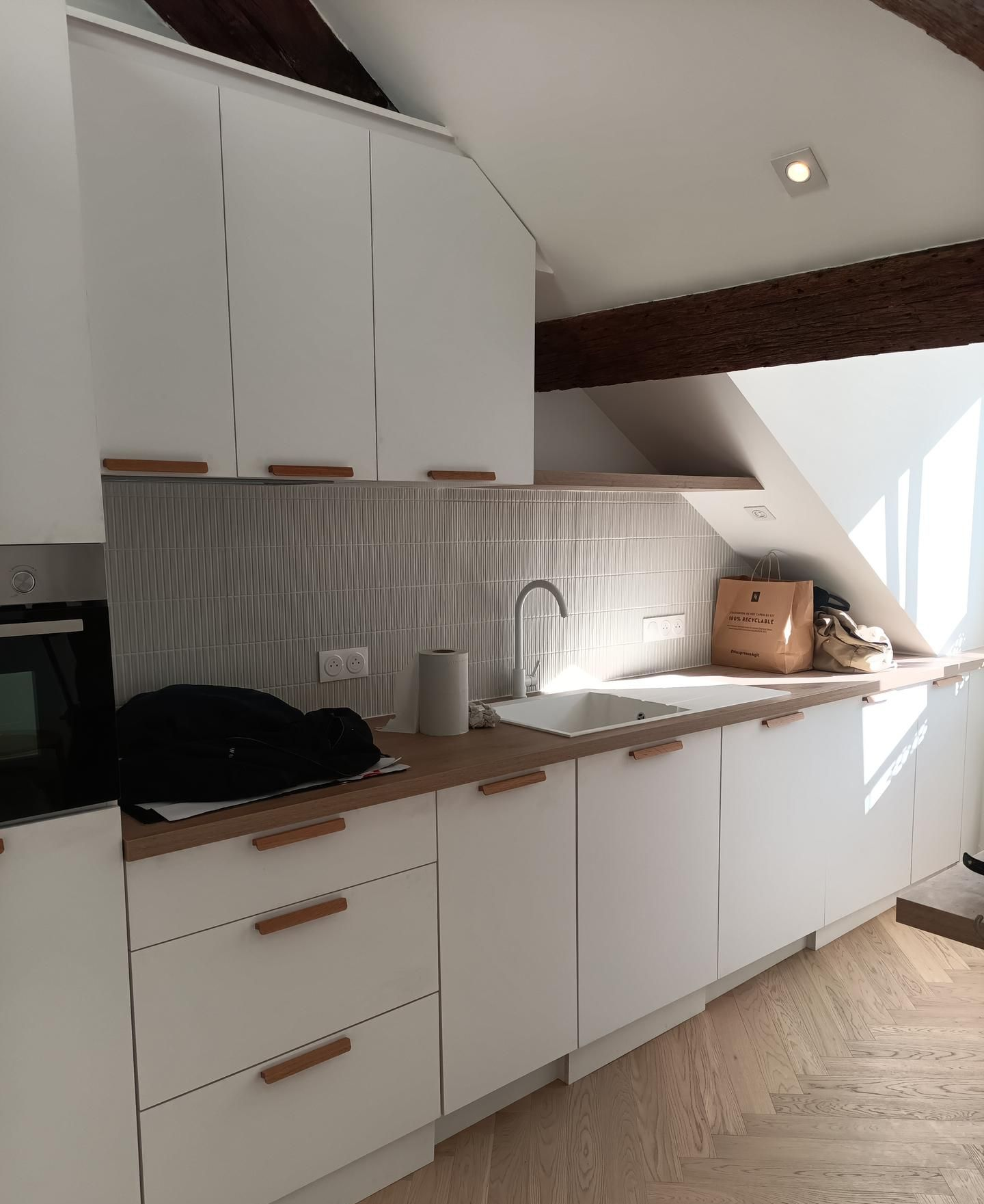 Cuisine équipée d'armoires blanches, d'un plan de travail en bois, d'un évier et d'une crédence carrelée, sous un plafond mansardé.