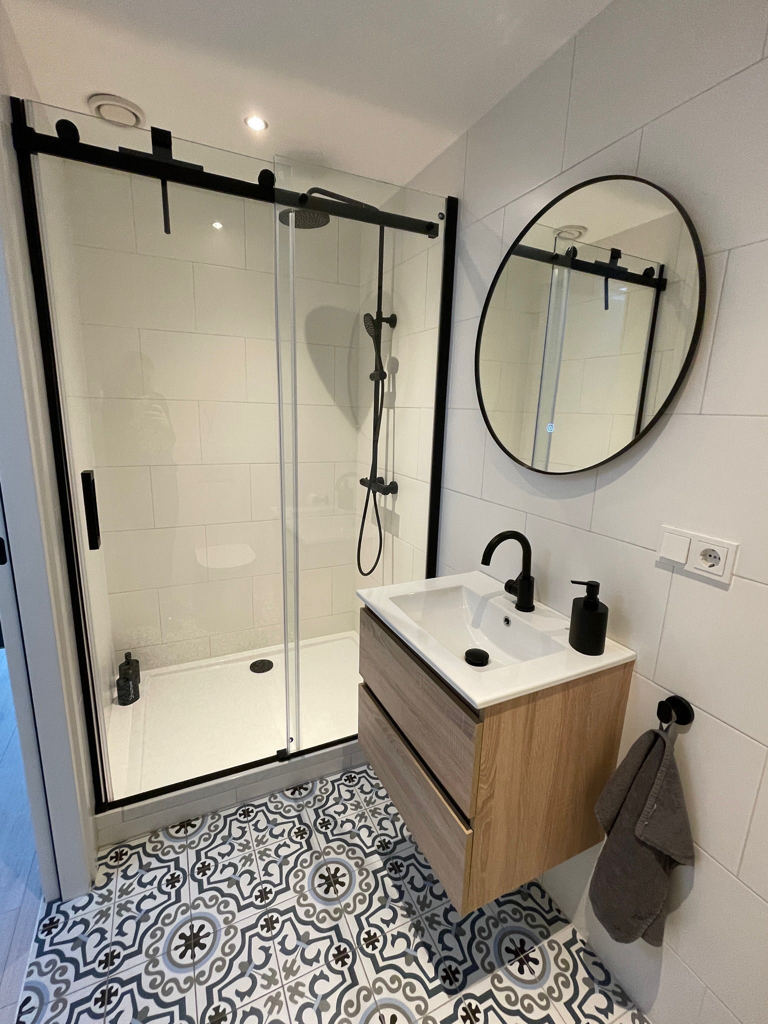 Petite salle de bains moderne avec douche à cadre noir, meuble-lavabo en bois, sol à motifs et miroir rond.