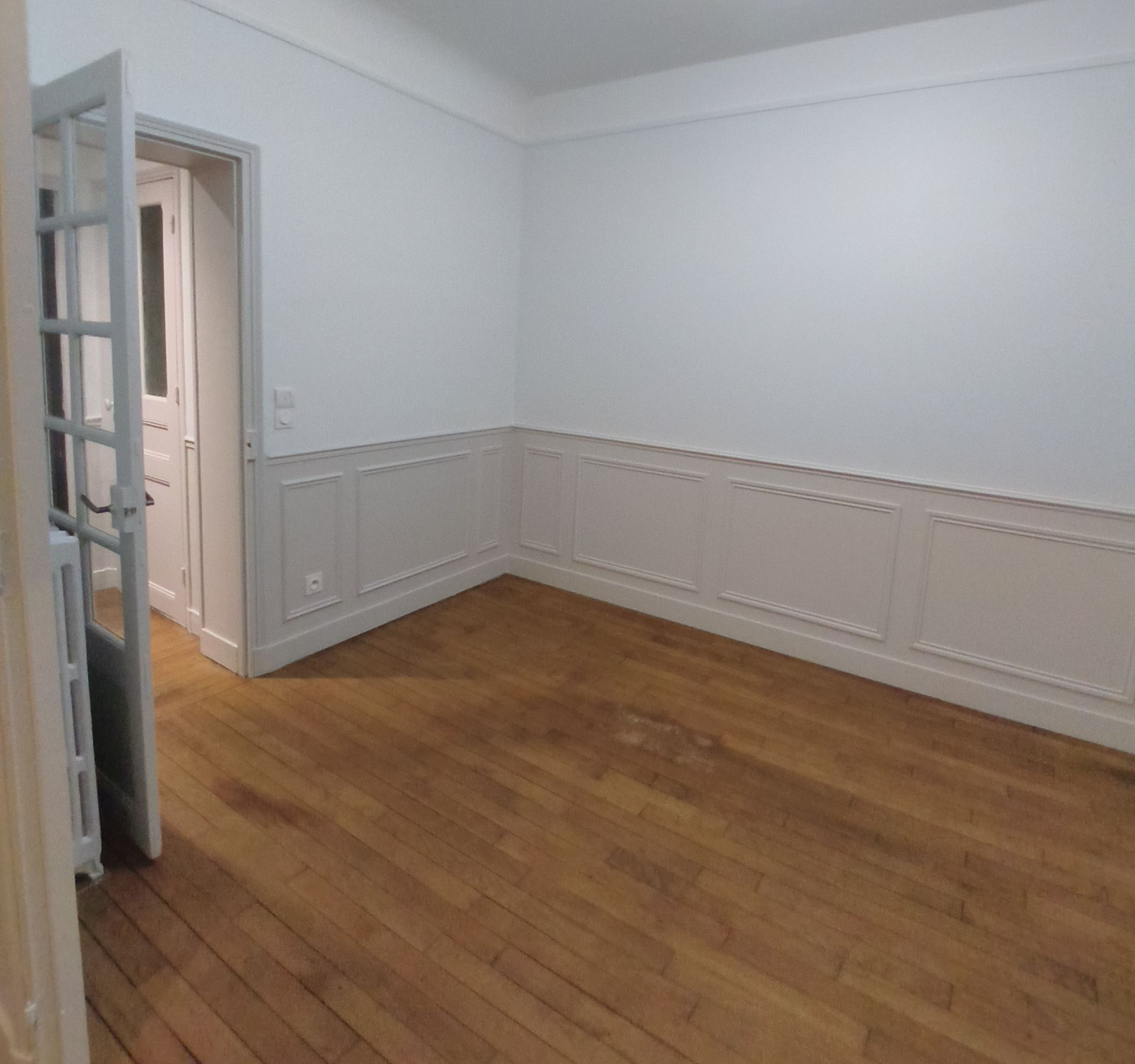 Pièce vide avec parquet et murs blancs. Une porte est entrouverte sur la gauche.