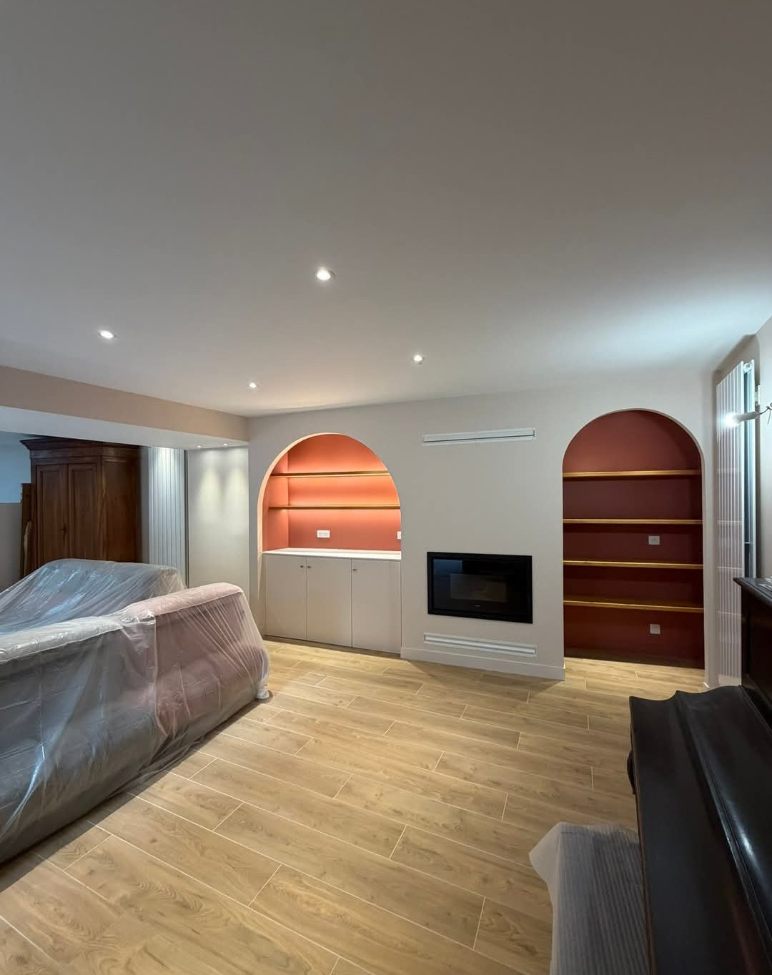 Salon avec parquet clair, étagères intégrées, cheminée, touches de brun rougeâtre et meubles recouverts de plastique.