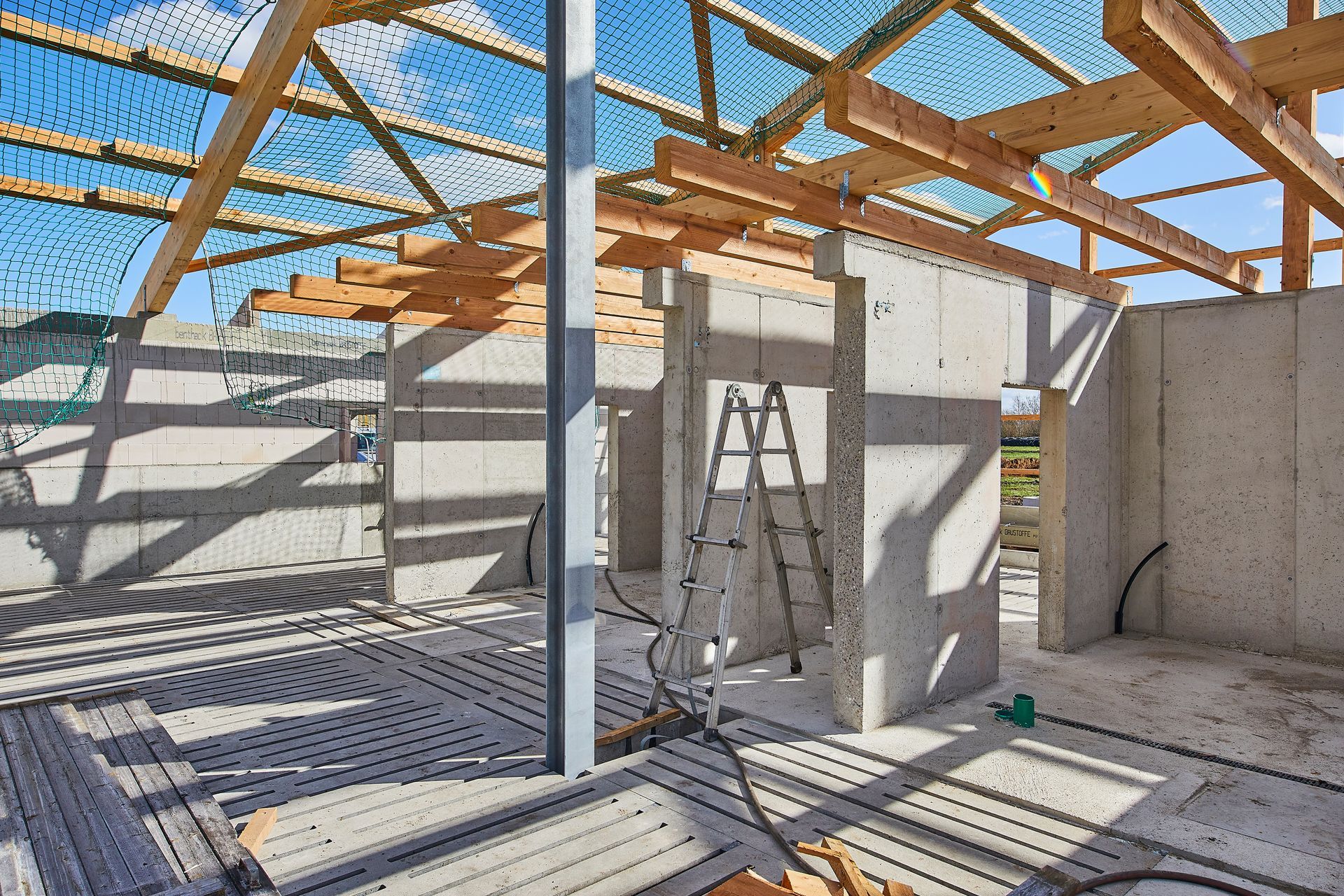 Innenansicht einer Baustelle mit Betonwänden, Holzbalken und einer Leiter unter blauem Himmel.