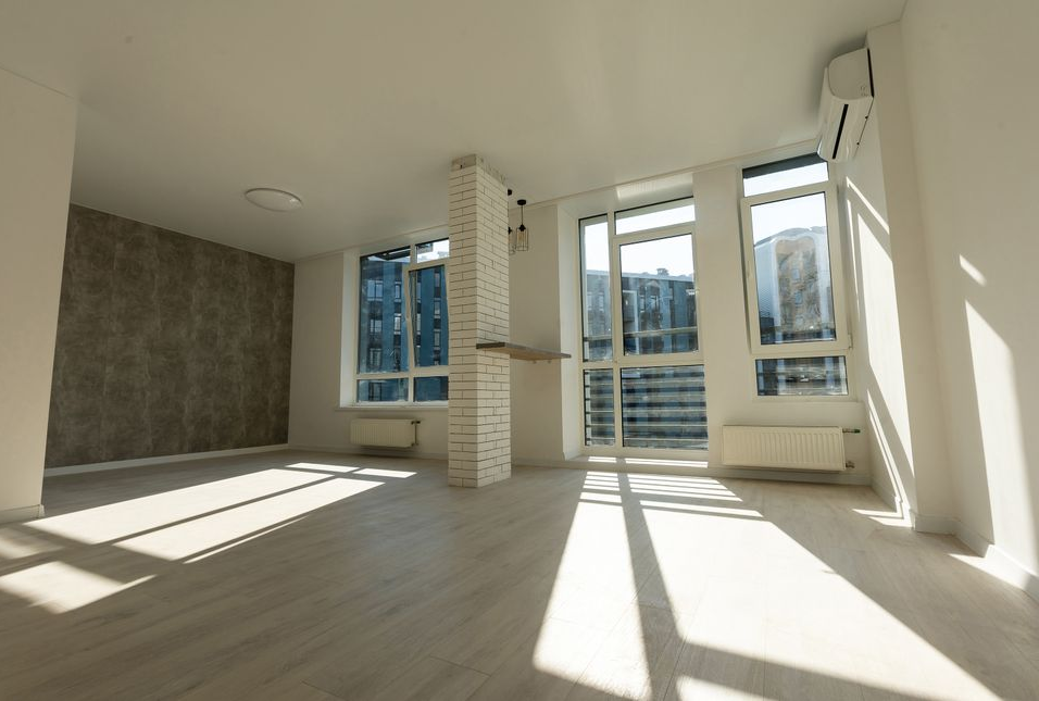 Leerer, sonnendurchfluteter Raum mit großen Fenstern, hellen Holzböden und einer strukturierten Akzentwand.