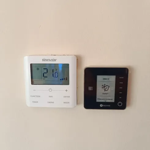Dos termostatos digitales de pared, uno blanco y otro negro, en una pared beige.
