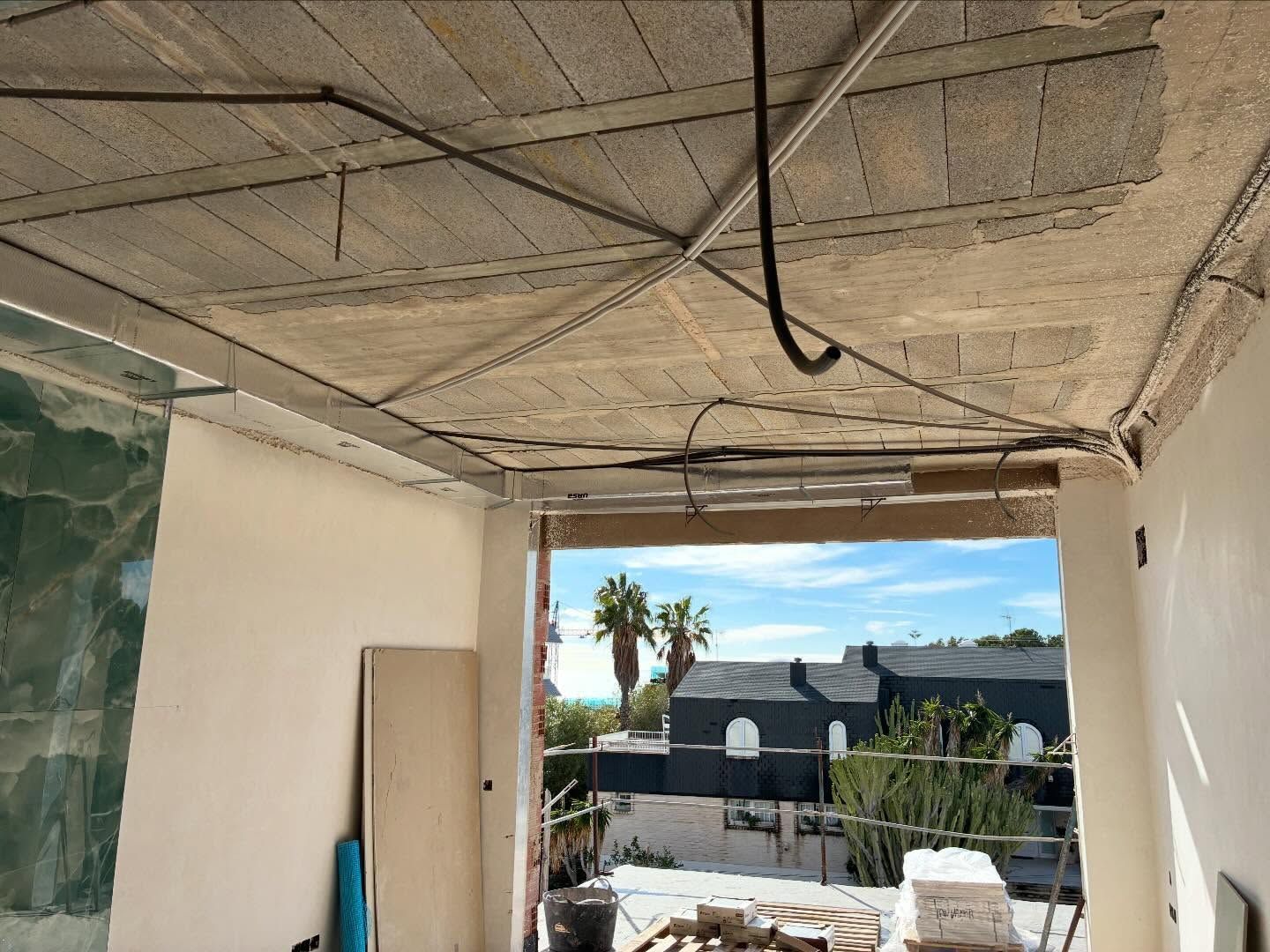 Interior de balcón sin terminar, con cables a la vista en el techo y vistas soleadas de tejados y palmeras