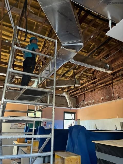 Un trabajador en una escalera instala conductos en el techo de una habitación sin terminar, entre vigas a la vista