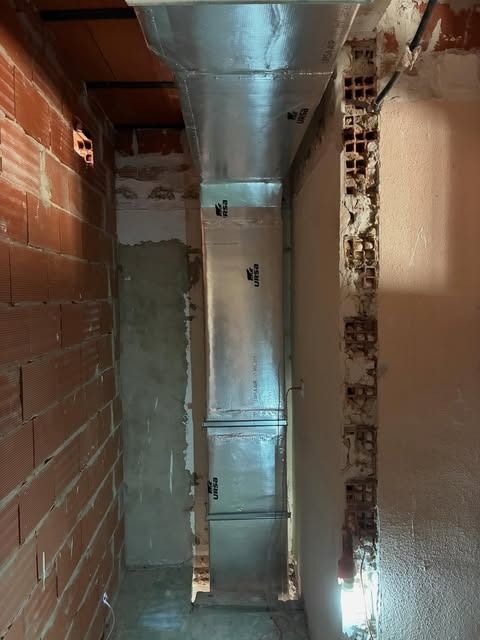 Pasillo estrecho sin terminar, con paredes de ladrillo visto y conductos metálicos en el techo