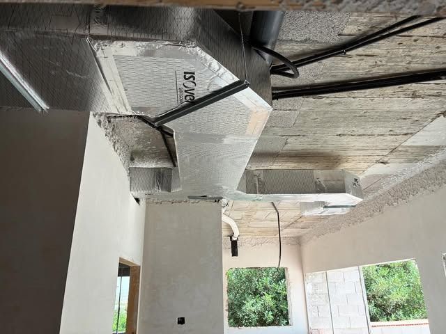 Habitación sin terminar, con conductos plateados a la vista y el techo abierto junto a una pared con ventanas