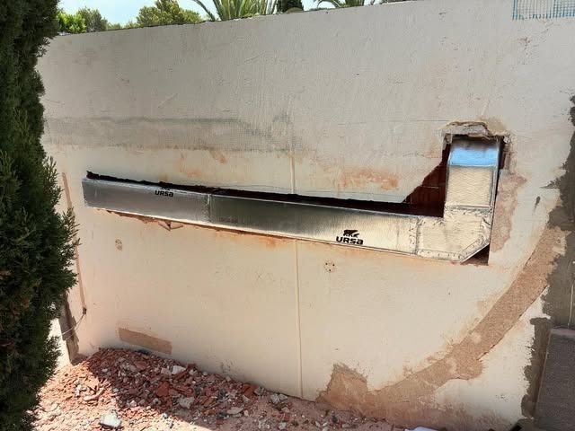Pared exterior dañada, con una larga abertura de ventilación o conducto y el yeso desmoronándose.