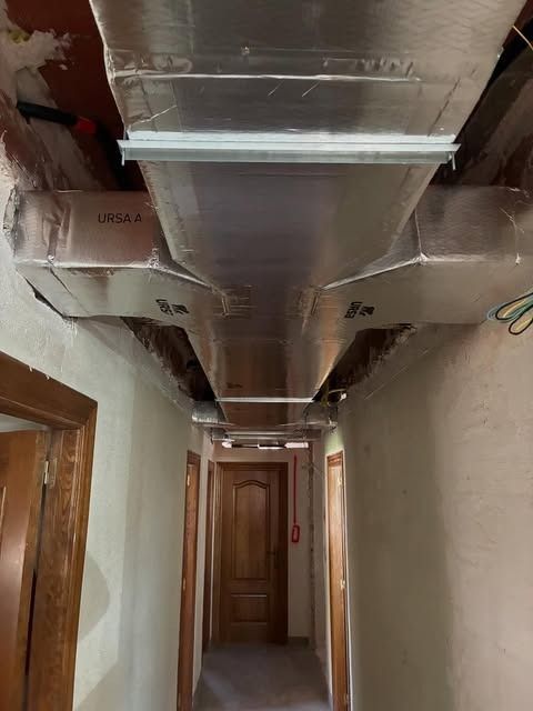 Pasillo estrecho con conductos de climatización a la vista a lo largo del techo, en un interior sin terminar.