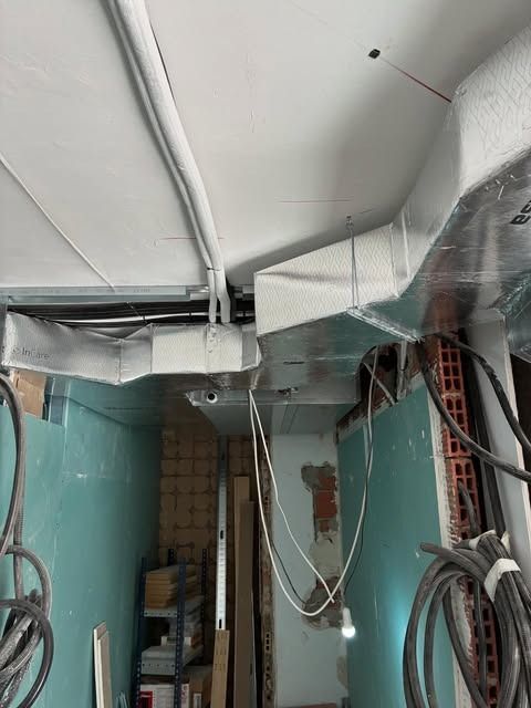 Conductos y cableado a la vista en una habitación estrecha sin terminar, con paredes verde azulado y techo blanco.