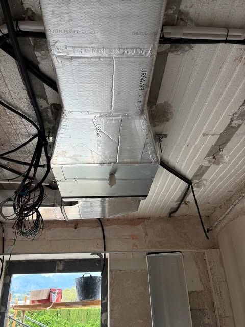 Conductos de climatización y cableado a la vista en el techo de una habitación con una ventana y paredes de ladrillo