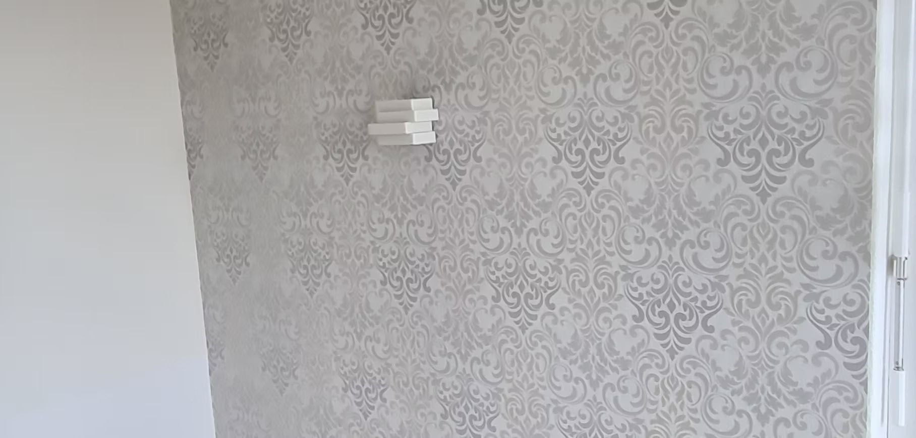Weisse Wand mit hellgrauer gemusterter Tapete und einer kleinen Wandleuchte im oberen Bereich (Mitte)