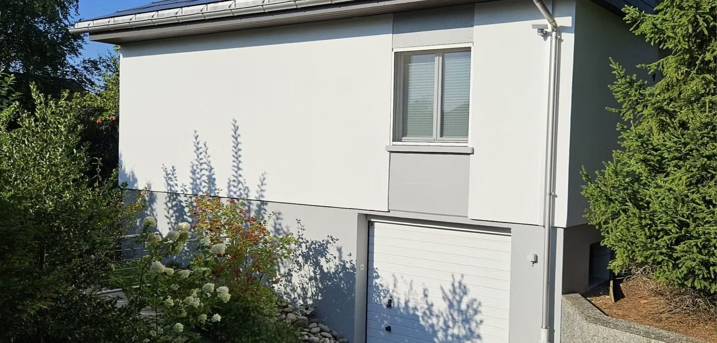 Weisses Haus mit kleinem Fenster, umzäuntem Garten und Bäumen auf beiden Seiten
